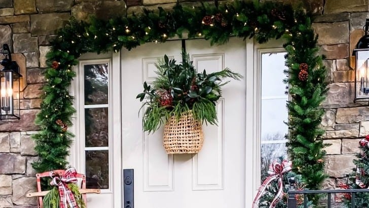 Cream front door on a stone house decorated for Christmas with garland and a basket of greenery.