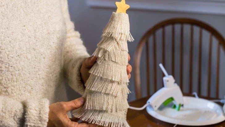 Woman holding a DIY tree decoration covered with rows of fringed burlap with a wood star on top.