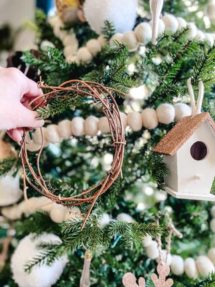 Hand hanging a small grapevine wreath ornament on a Christmas tree decorated with felted ball garland.
