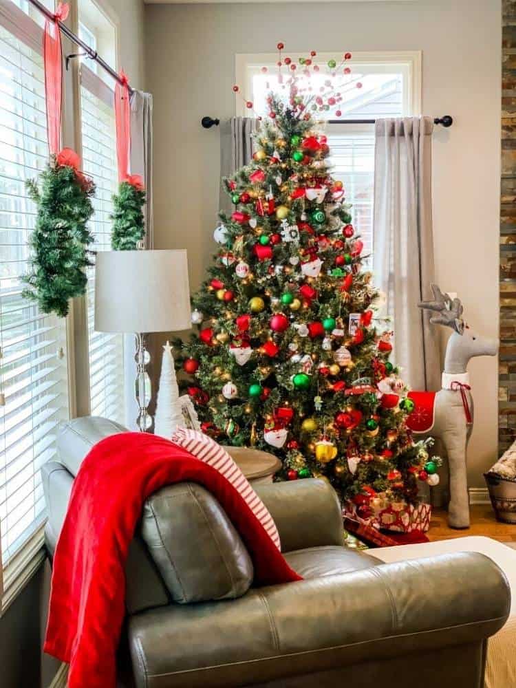 Traditional red and green ornaments fill a Christmas tree in a family room with wreaths on the windows.