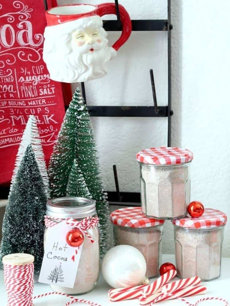 Hot cocoa bar with a Santa mug and jars of hot cocoa mix with red and white checked lids and bottle brush trees.