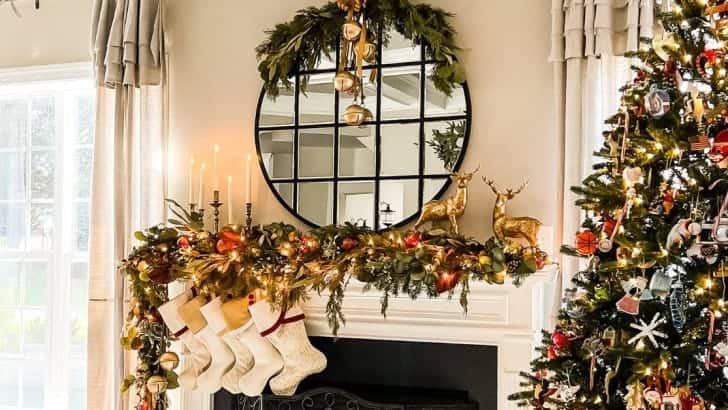 Gas fireplace with asymmetrical Christmas garland, stockings, round mirror with greenery and gold reindeer.