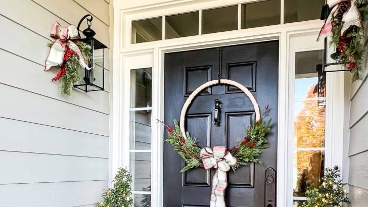 Black front door with transom and sidelights decorated for Christmas with a hoop wreath, greenery, and plaid ribbon.