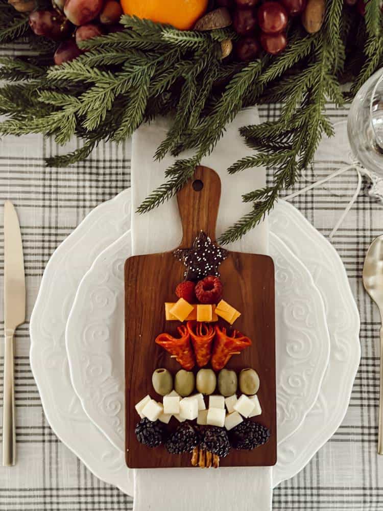 Mini board with a charcuterie Christmas tree on top of a place setting at a table with greenery.