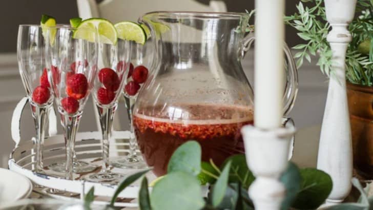 Pitcher of raspberry prosecco cocktail with champagne glasses filled with fresh raspberries on a table.