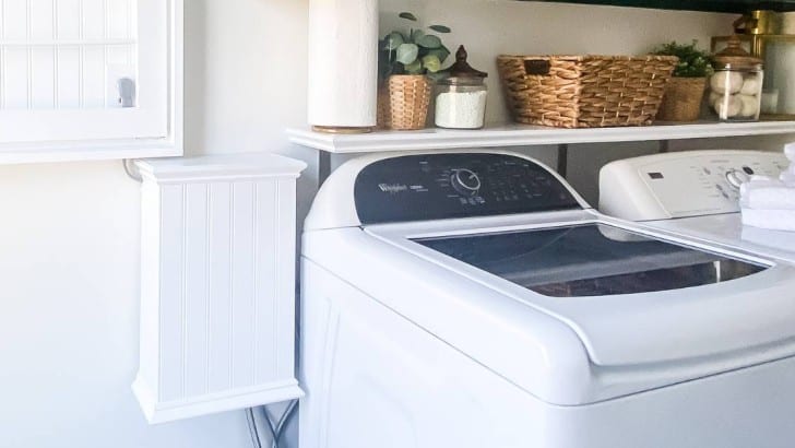 Laundry room with top load washer and a shelf above. Washer hookups are covered with a custom beadboard box.