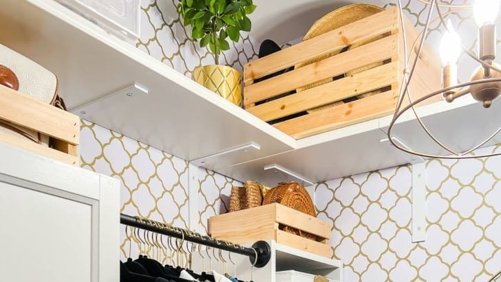 Shelf at top of a wallpapered closet hung with white steel L brackets with wood crates holding purses and hats.
