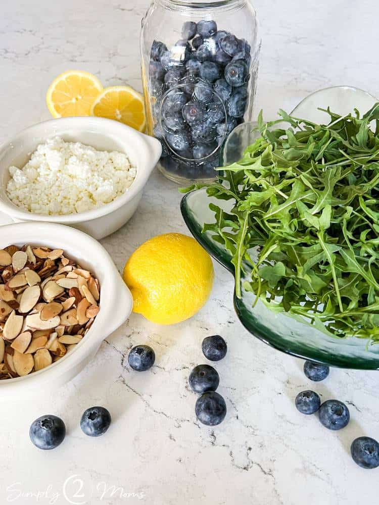 Glass salad bowl filled with baby arugula with a mason jar of fresh blueberries, lemons, bowl of goat cheese and bowl of toasted almonds.