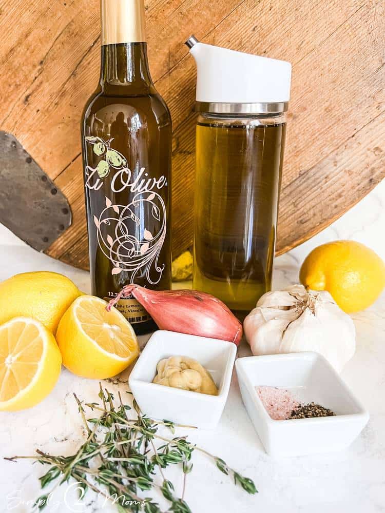 Bottles of white lemon balsamic vinegar and olive oil surrounded by lemons, shallots, head of garlic, fresh thyme and seasonings.
