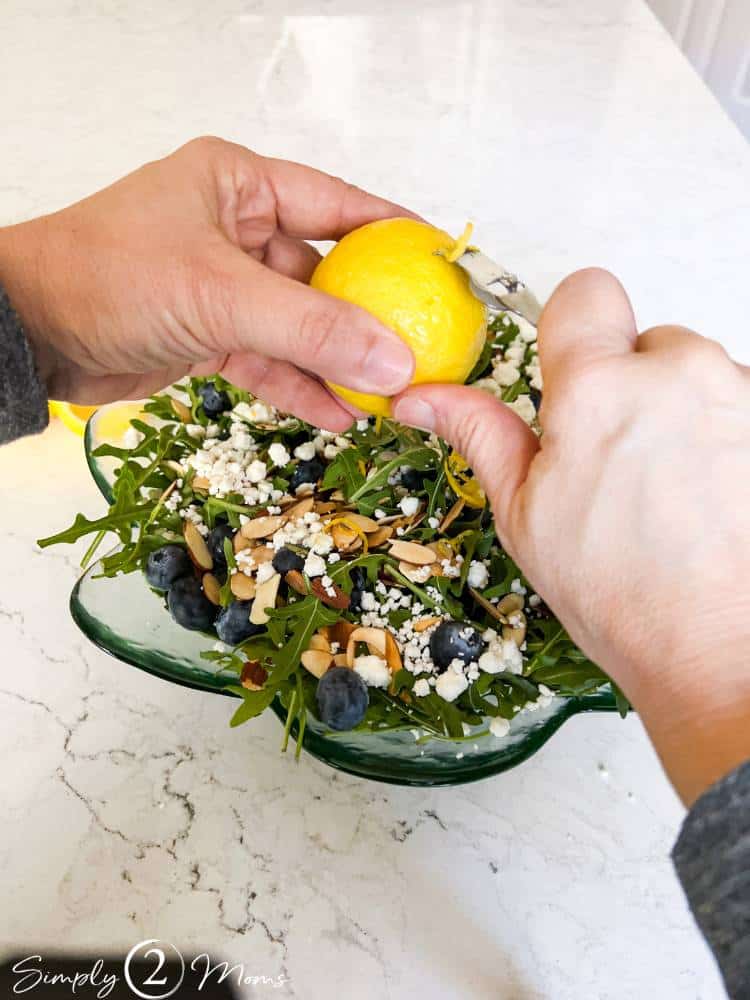 Woman using a zester tool to add fresh lemon zest to a blueberry salad.