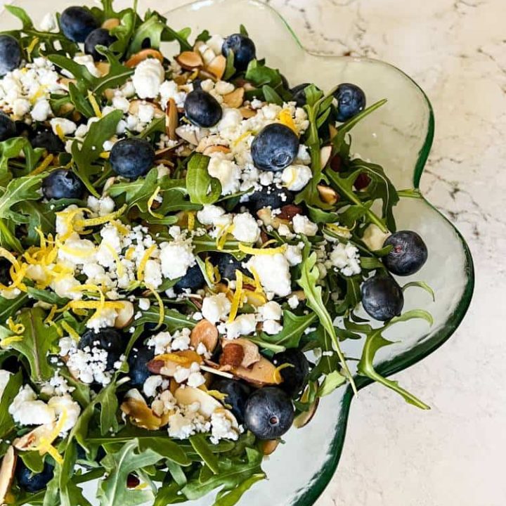 Serving bowl of arugula salad with fresh blueberries, toasted almonds, goat cheese, and lemon zest.