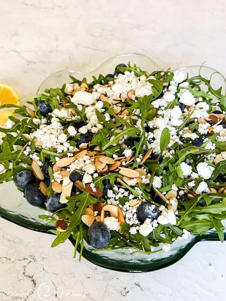 Serving bowl of arugula salad with fresh blueberries, toasted almonds, goat cheese, and lemon zest.