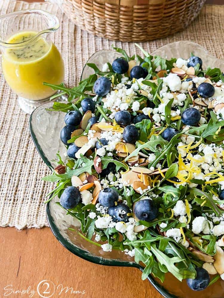 Serving bowl of blueberry arugula salad topped with goat cheese and a small pitcher of lemon vinaigrette.