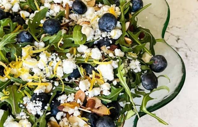 Serving bowl of arugula salad with fresh blueberries, toasted almonds, goat cheese, and lemon zest.