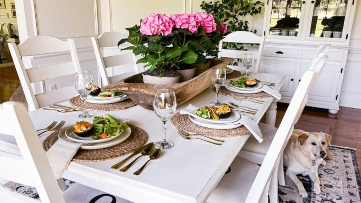 White dining table with deep pink hydrangeas in a trencher bowl set for Mother's Day Lunch with baked avocados on the plates.