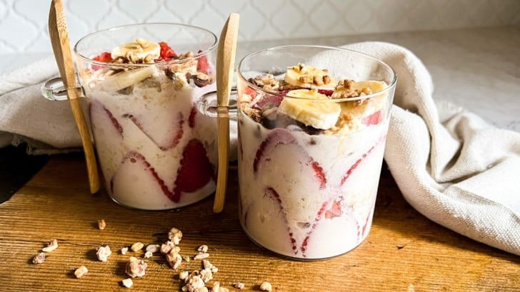 Two glass containers with wood spoons filled with healthy strawberry banana overnight oats topped with granola.