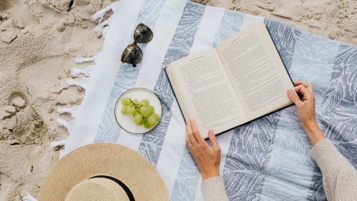 Fringed beach blanket on the sand with a woman's hands holding an open book beside a straw hat and a plate of grapes.
