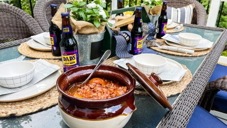 Crock of baked beans on an outdoor table set with plates and bowls and a Father's Day toolbox centerpiece.