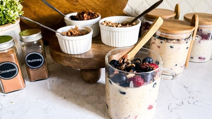 Glass jars with bamboo lids filled with mixed berry overnight oats with ramekins of toppings and jars of cinnamon and nutmeg.
