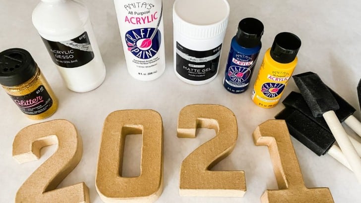 Containers of acrylic gesso, acrylic paints, matte gel medium, and foam brushes with papier mache numbers.