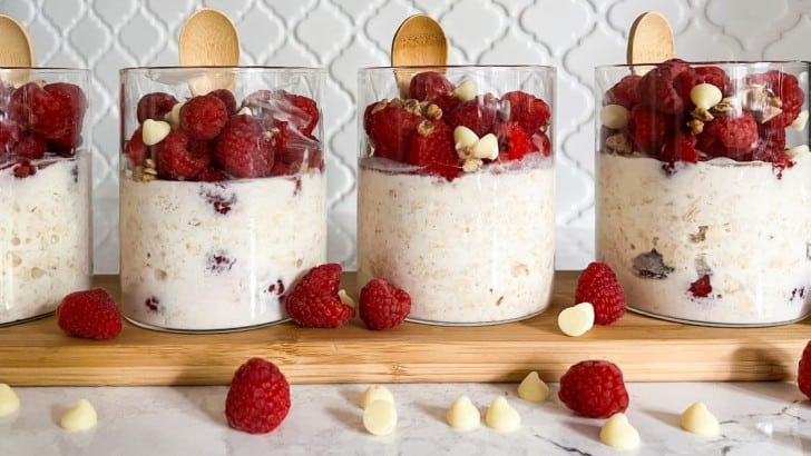 Four glass jars with attached bamboo spoons filled with white chocolate raspberry overnight oats with fresh raspberries surrounding them.