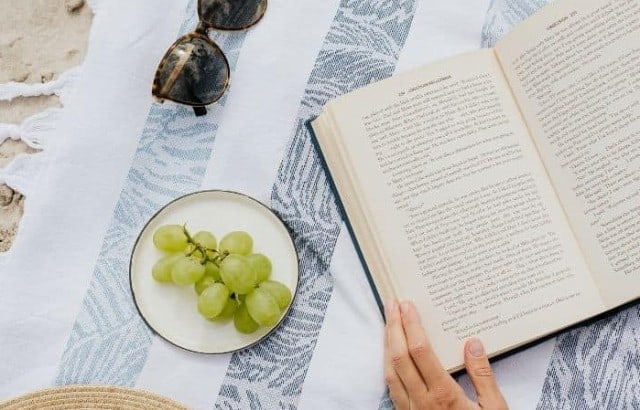 Fringed beach blanket on the sand with a woman's hands holding an open book beside a straw hat, sunglasses, and a plate of grapes.