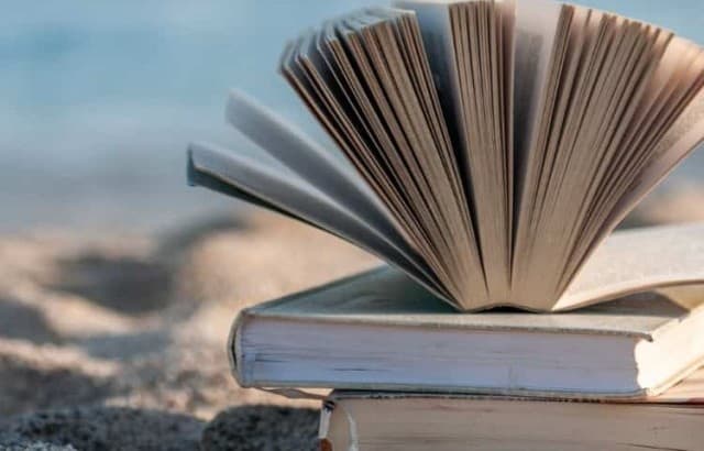 Stack of three books on a sandy beach with the water out of focus behind them.