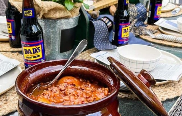 Crock of baked beans on an outdoor table set with plates and bowls and a Father's Day toolbox centerpiece.