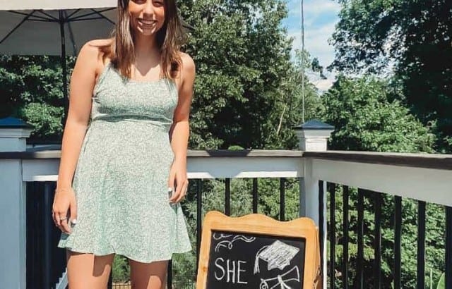 graduate standing on deck outside next to chalkboard sign