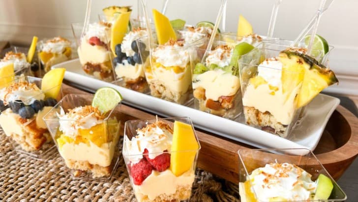 Mini plastic cups of keto tropical trifles with a variety of diced fruit on a platter with wood base.