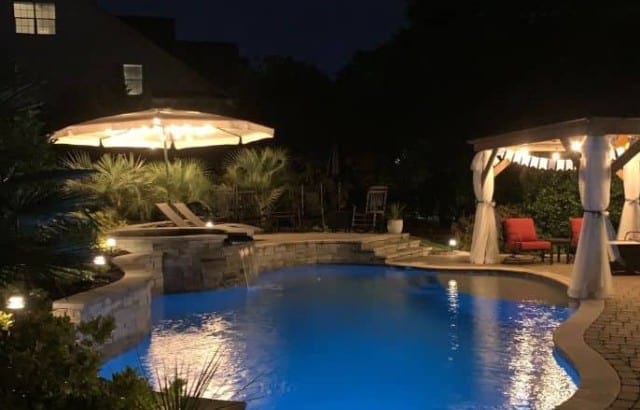 Custom inground pool at night with umbrella and gazebo lit with stringlights.