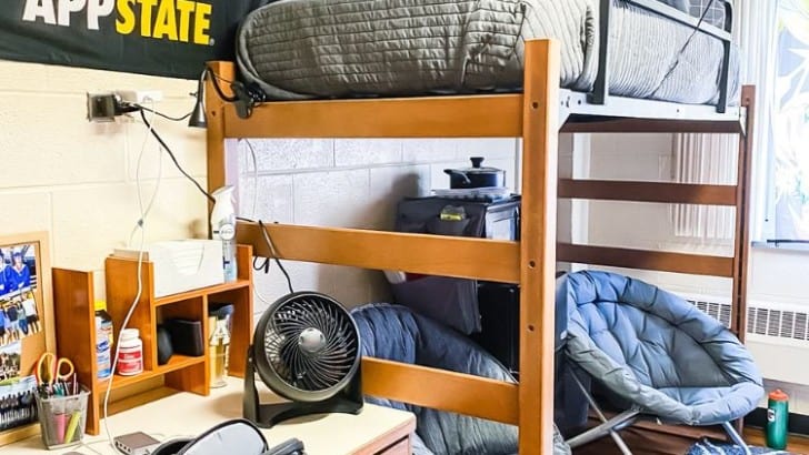 Guy's dorm room with lofted bed, folding saucer seats, desk, and college flag on the wall.