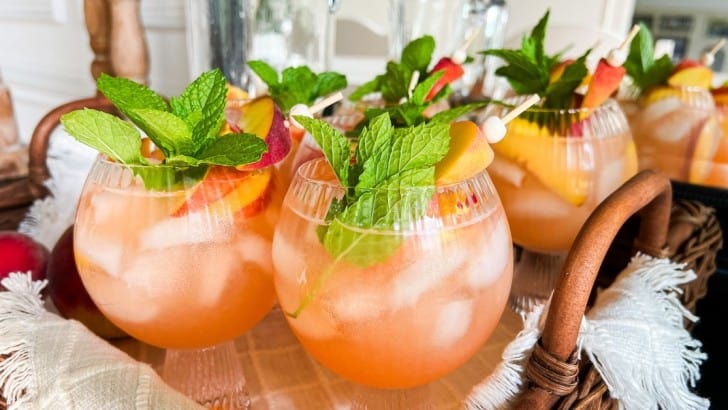 Round goblets on a wicker serving tray filled with peach sangria garnished with sliced peaches and mint.