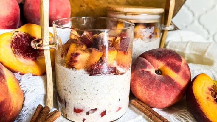 Jar of peach cobbler overnight oats surrounded by fresh peaches and sticks of cinnamon.
