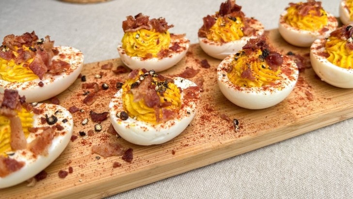 Wood serving board with southern deviled eggs topped with bacon, cracked black peppercorns, and smoked paprika.