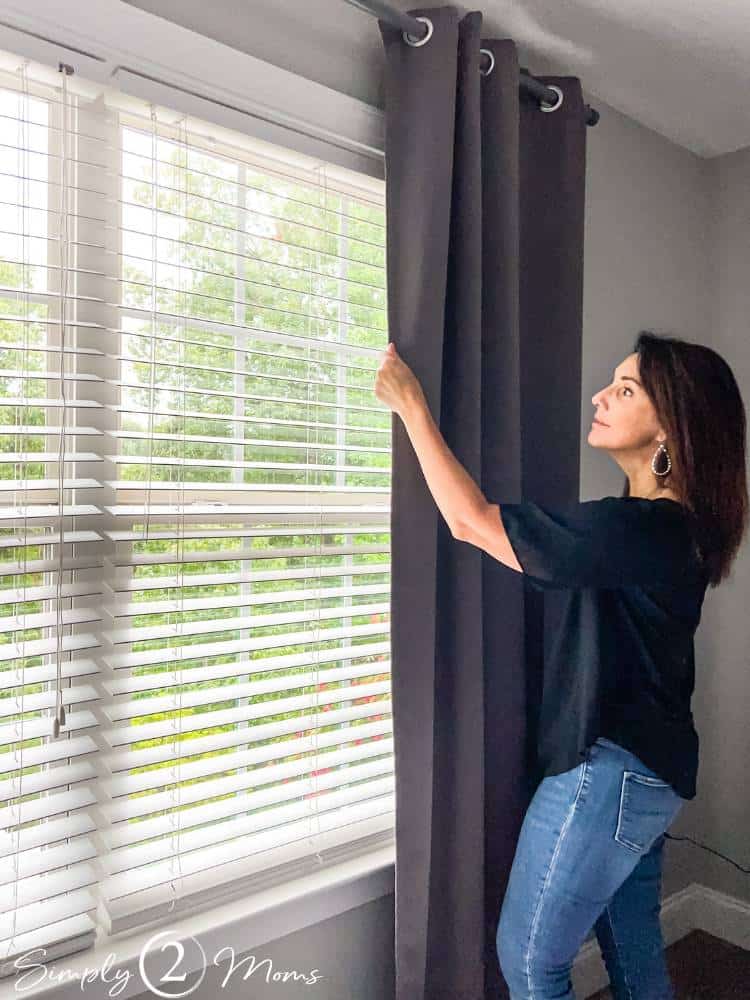 AnnMarie pulling curtain open hanging on window with white faux wood blinds