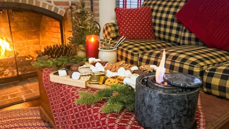 Wood board filled with everything to make Christmas s'mores set up on a table with an indoor fire pit beside a lit fireplace.