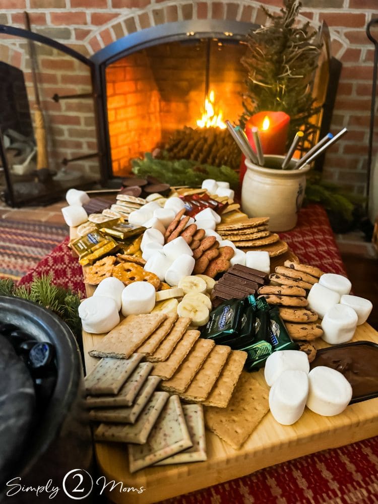 Wood board filled with ingredients to make s'mores in a room with a lit fireplace.