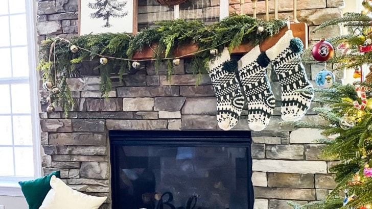 Stone fireplace with wood mantel decorated for Christmas with greenery, bells, and green and white stockings.
