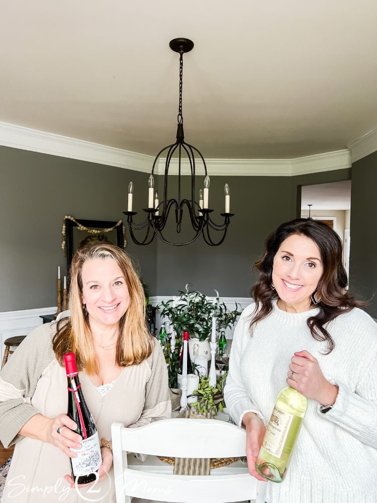 Two women standing in front of dining table holding bottles of wine for dinner party