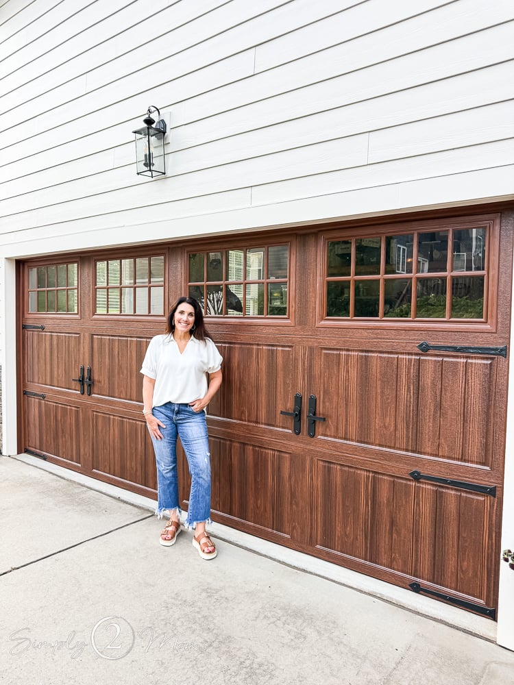 Woman standing in front of a faux wood double garage door with windows and iron hardware.