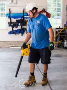 Installer wearing hard hat, googles, ear buds and gloves using a blower to clean up after a garage door replacement.