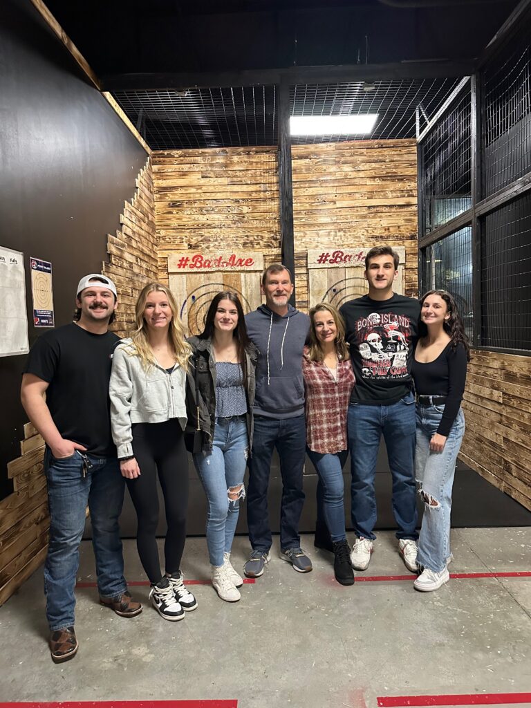 Family group smiling together at an axe throwing venue, enjoying a fun and adventurous outing with young adults.