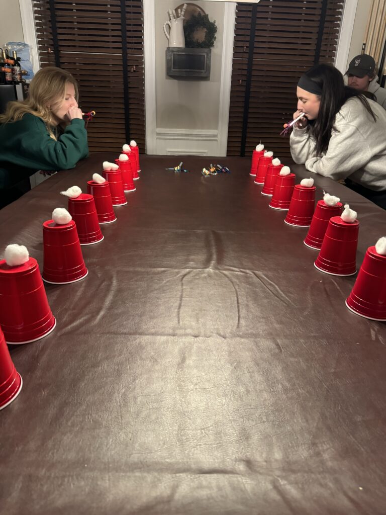 Two young women competing in a Christmas game by blowing cotton balls off red cups with party blowers.