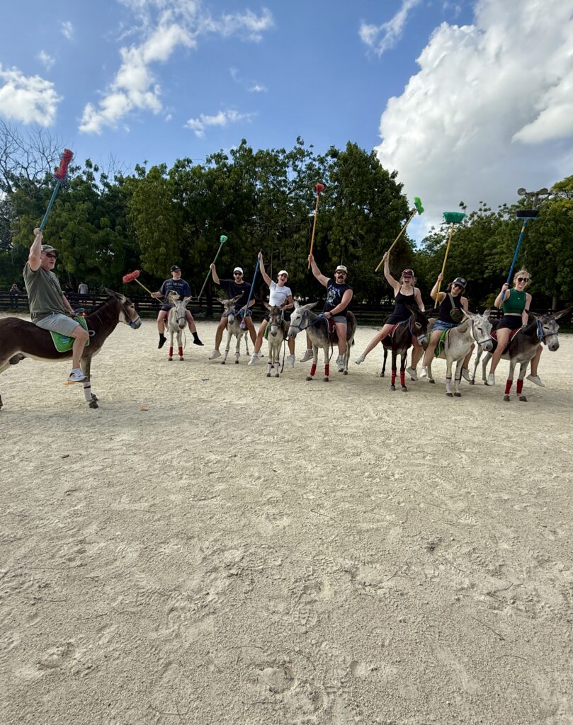 Group of family and friends riding donkeys with broomsticks raised, playing a lively game of donkey polo on vacation in the Dominican Republic.