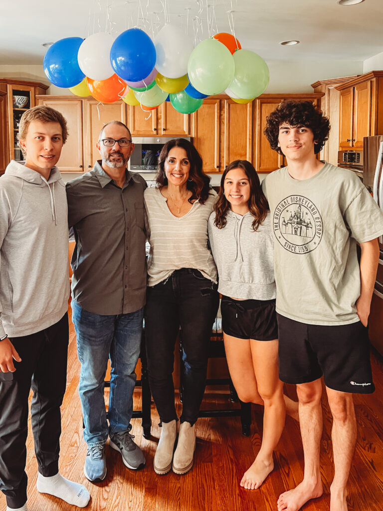 Family standing together in the kitchen under colorful balloons, celebrating a birthday with young adult children at home.