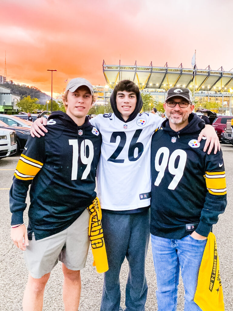 Dad and two sons wearing Steelers jerseys at a football game with stadium in the background, sharing a memorable sports gift experience.