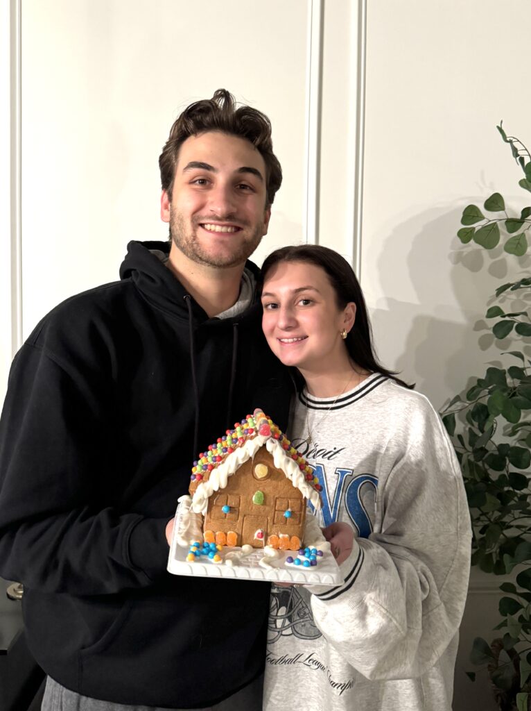 Smiling couple holding a decorated gingerbread house covered with colorful candies and icing during a family Thanksgiving contest.