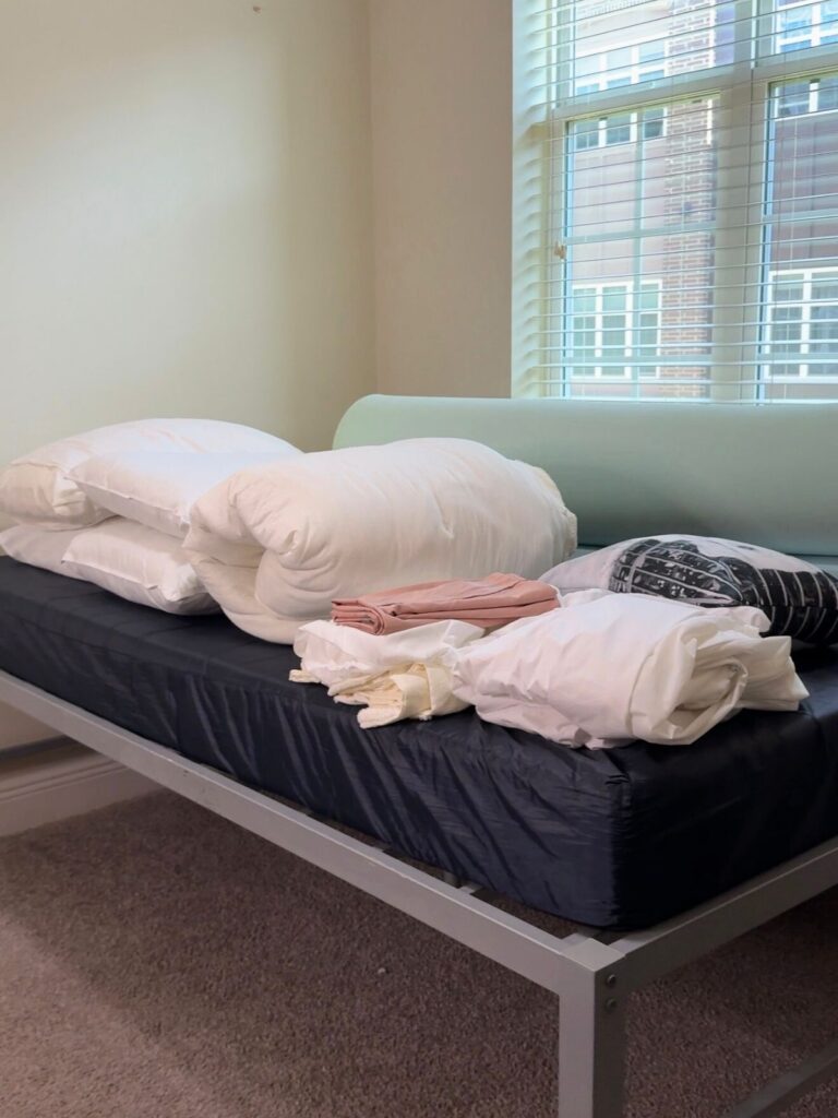 Dorm bed with white bedding, black mattress topper, and folded sheets on metal frame