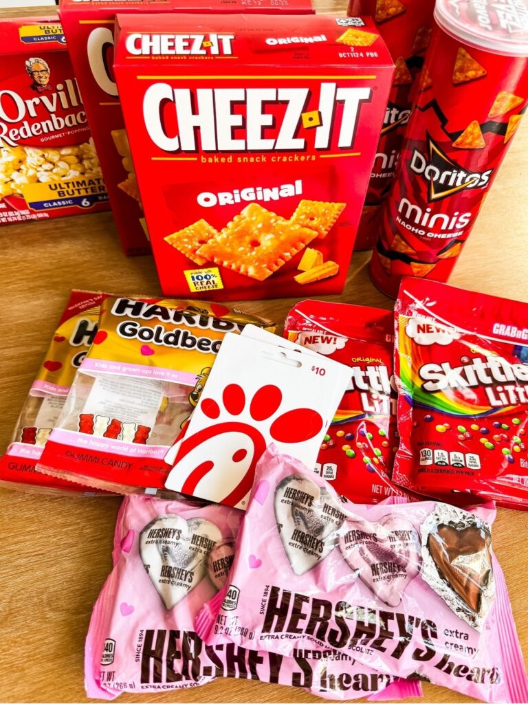 Red-themed college care package snacks including Cheez-It crackers, popcorn, candy, chocolate hearts, and a gift card laid out on a table